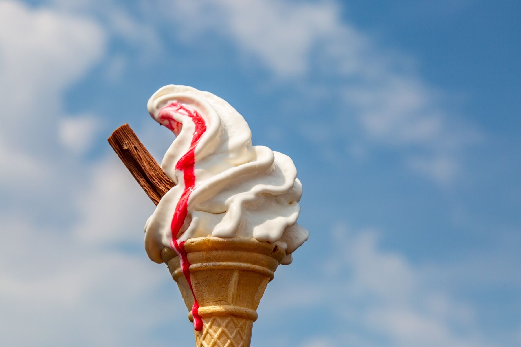 An ice cream cone with whippy ice cream, a flake and red sauce against a blue sky