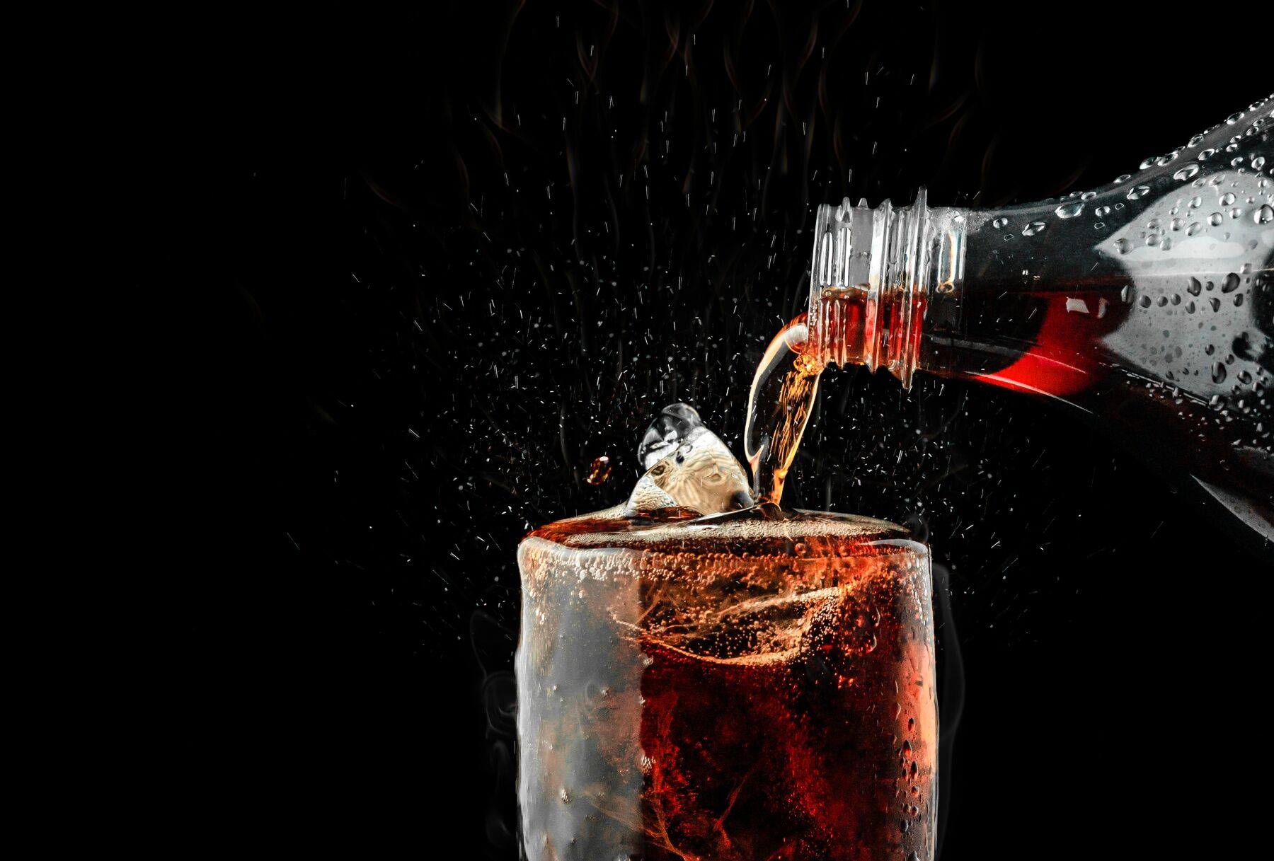 Soda being poured from a bottle into a glass full of ice cubes, against a black background