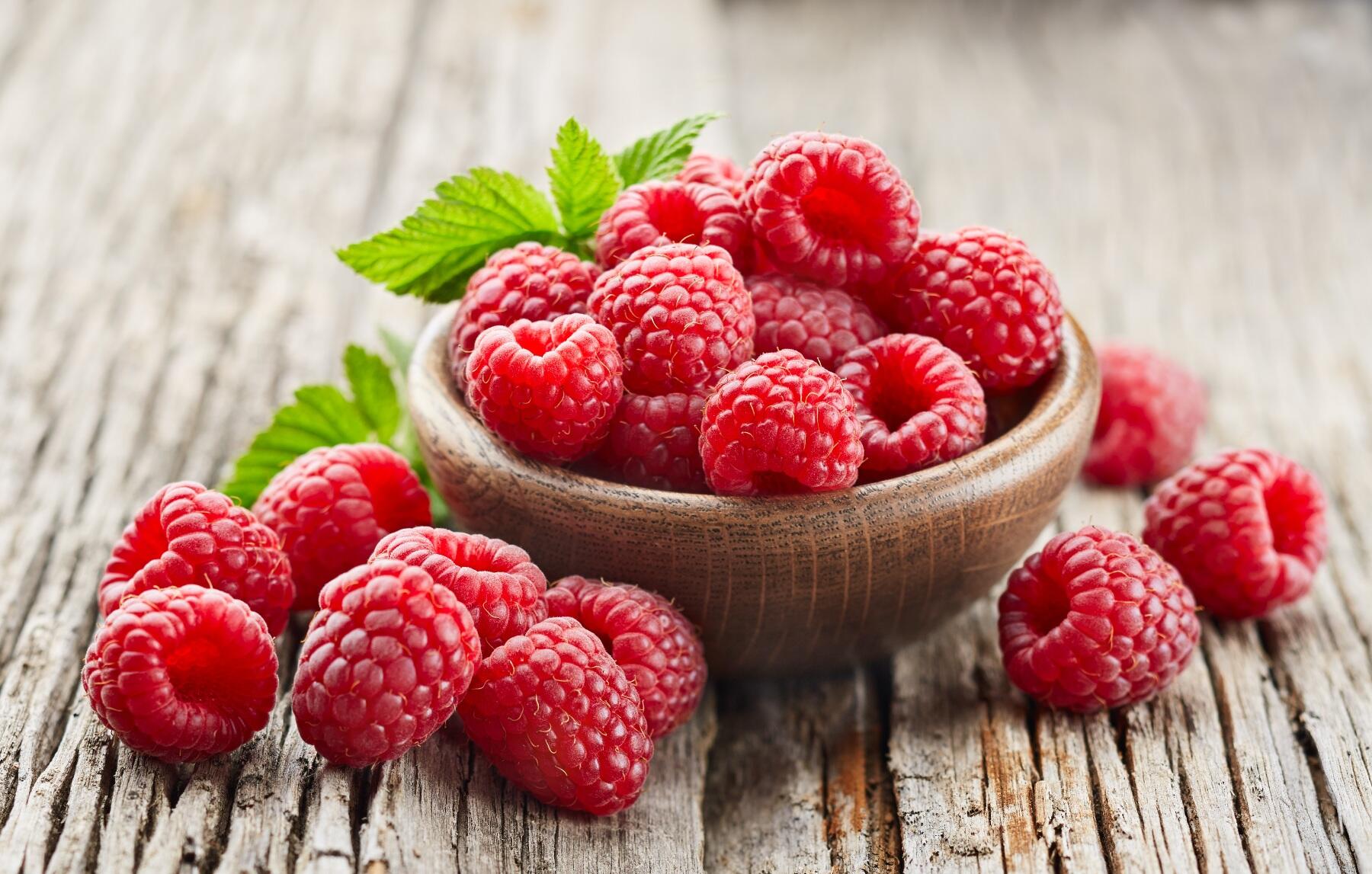 Raspberries in a little wooden bowl on a wooden table and spilling out of the bowl