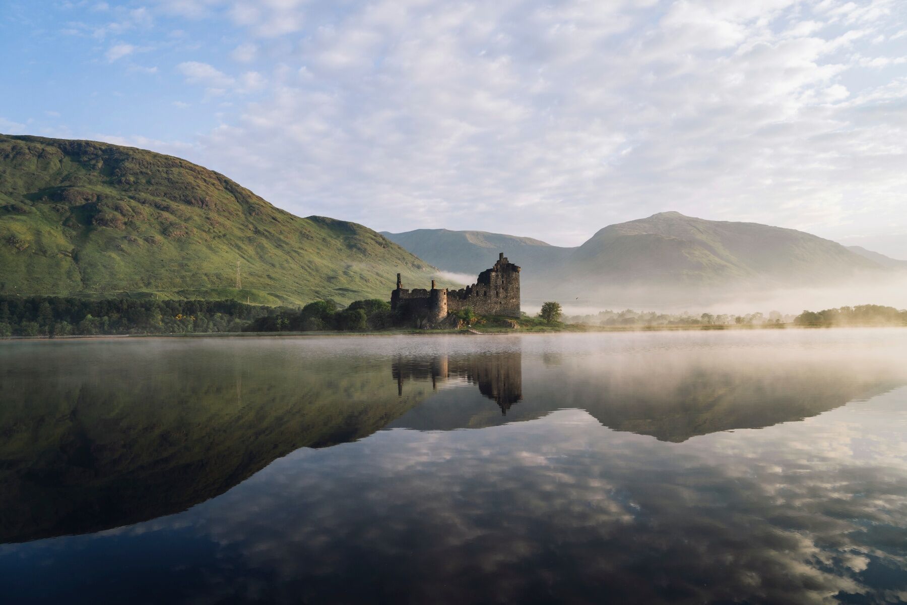 A castle in scotland, shrouded in mist next to a lake or river