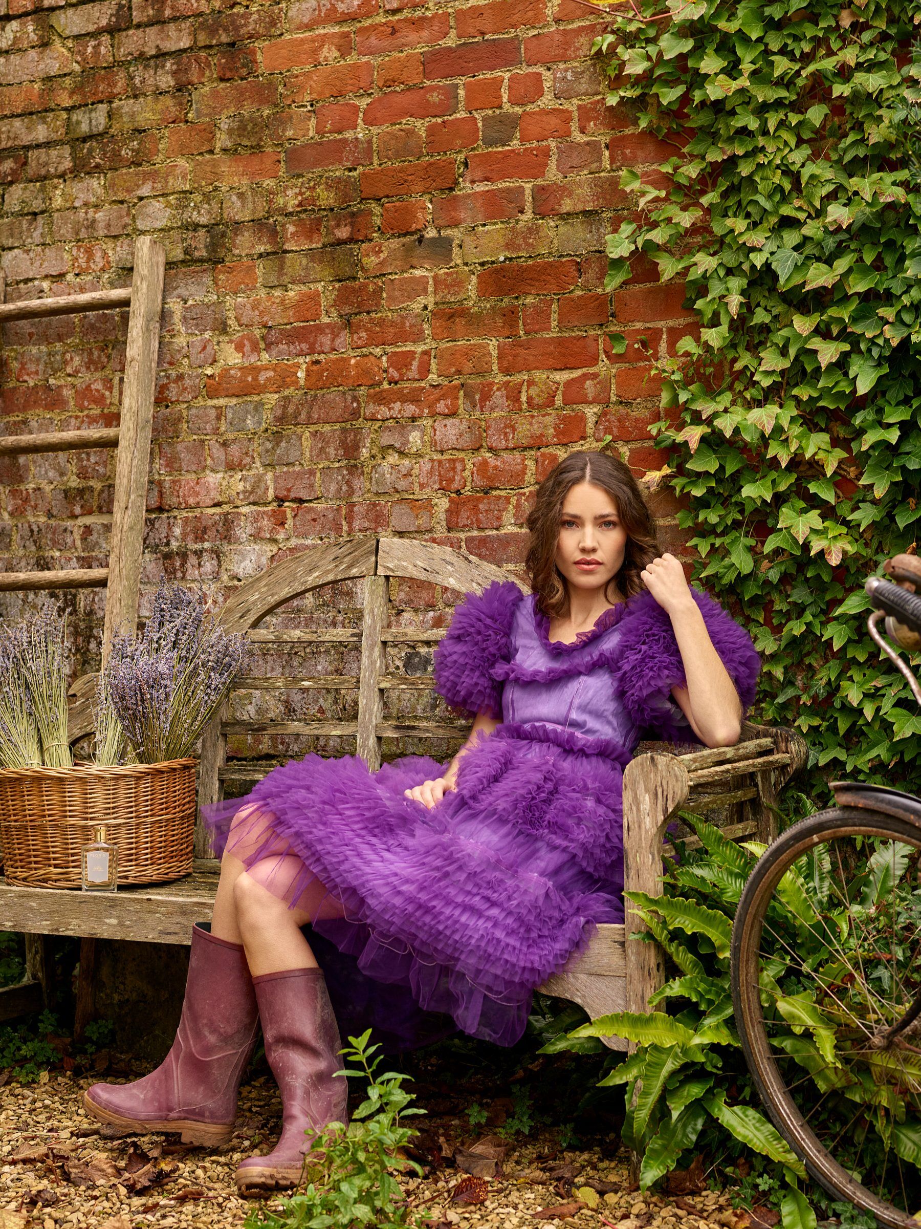 A woman in a purple frilly dress sat on a bench wearing wellies