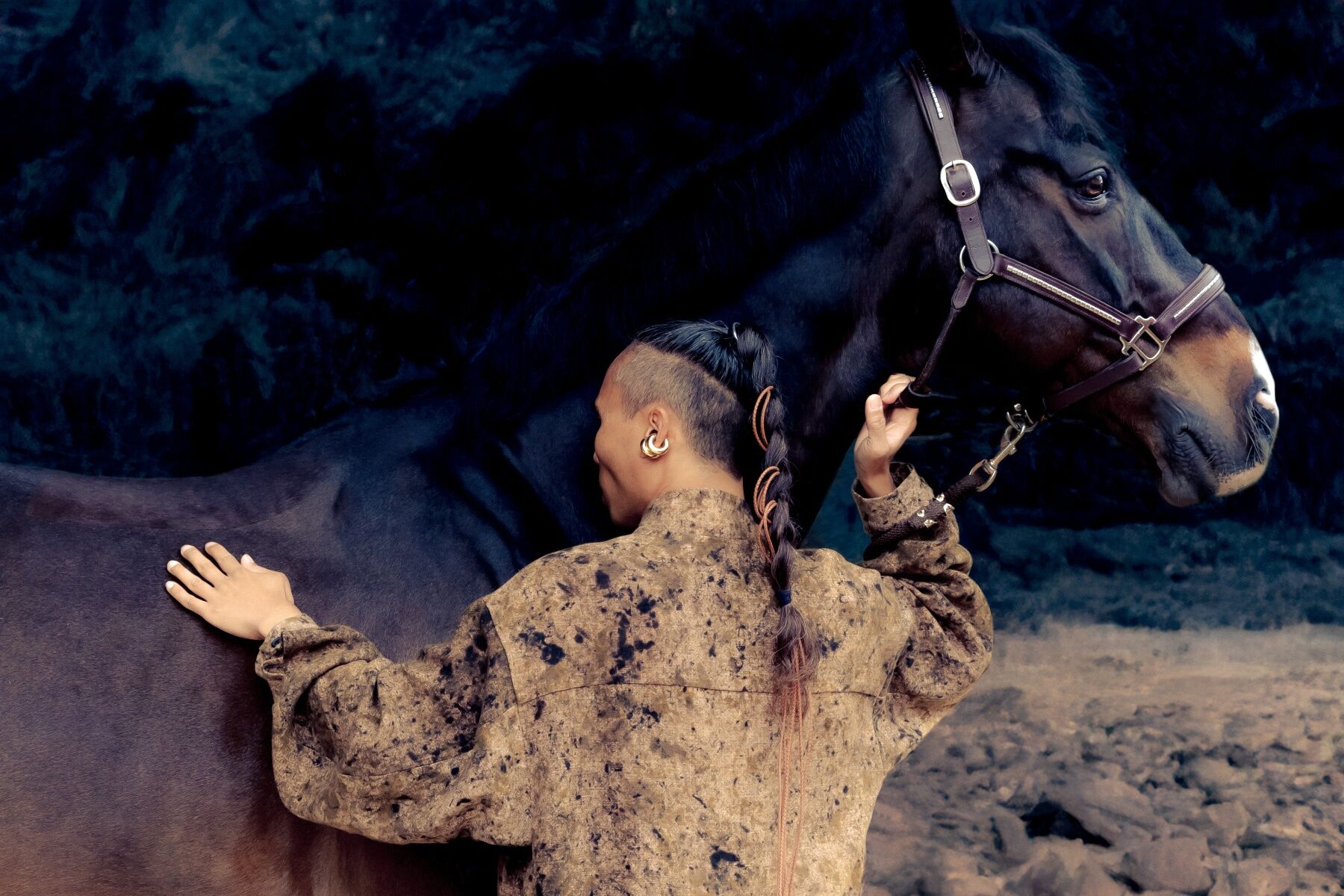 A man in a leather jerkin with a long braid and his back to the camera stroking an immense black horse