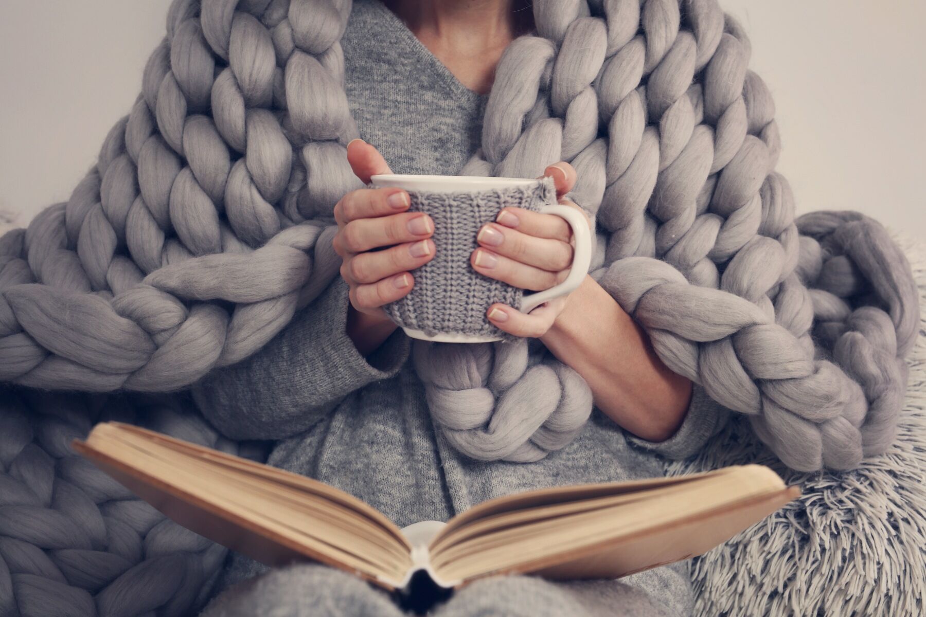 A person with a mug of tea and a large book sat cross legged under a huge blanket