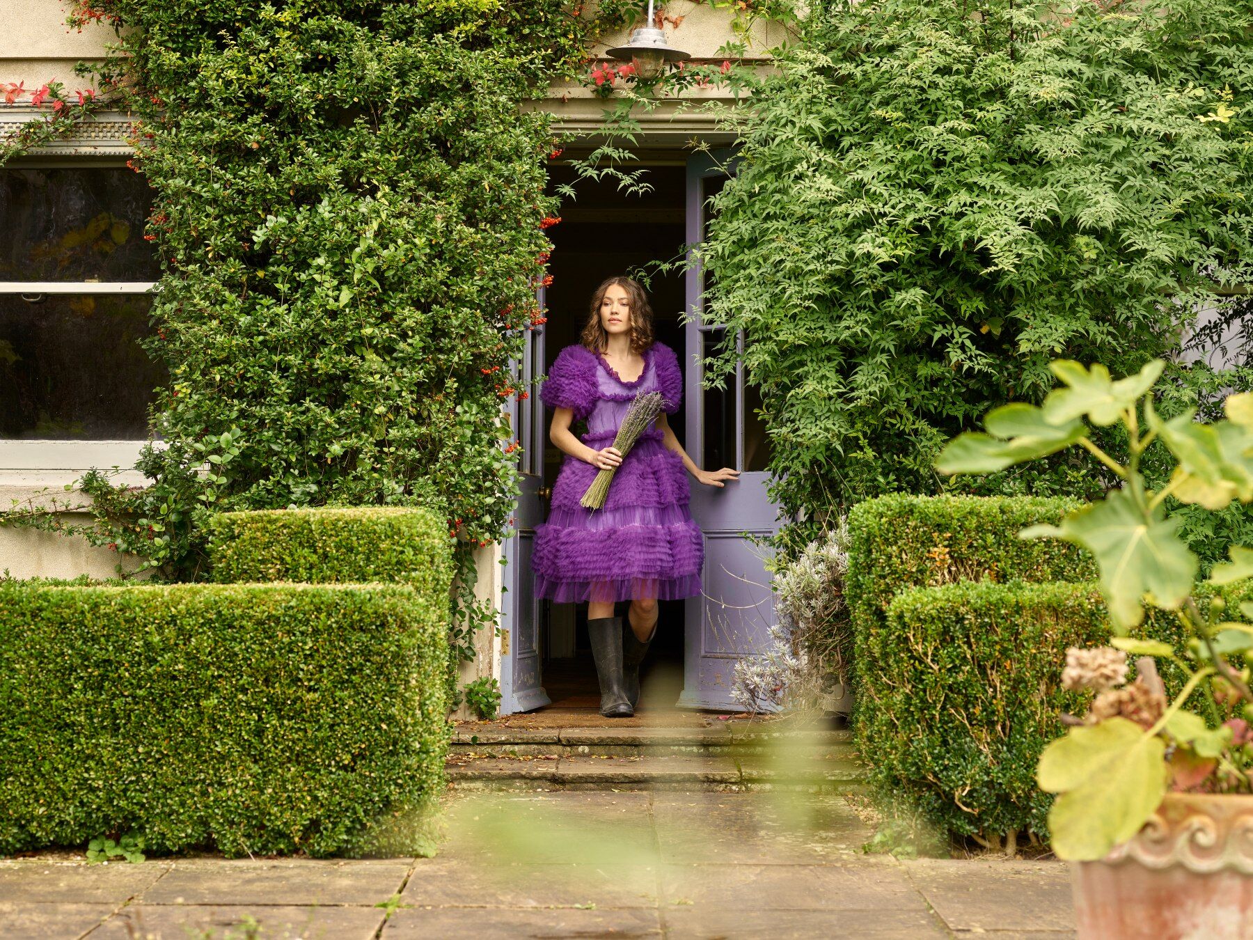 A woman in a purple dress and wellies coming out of a massive country house