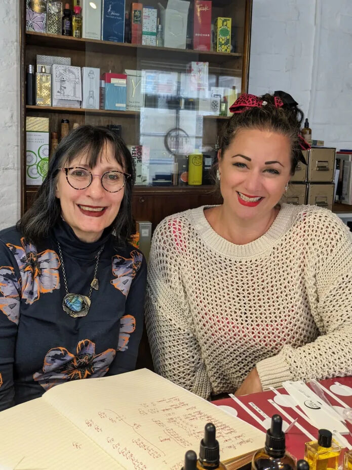 Sarah McCartney (left) with Tempest Rose (right) sat in front of a shelf full of perfumes, more materials are scattered on the table in front of them