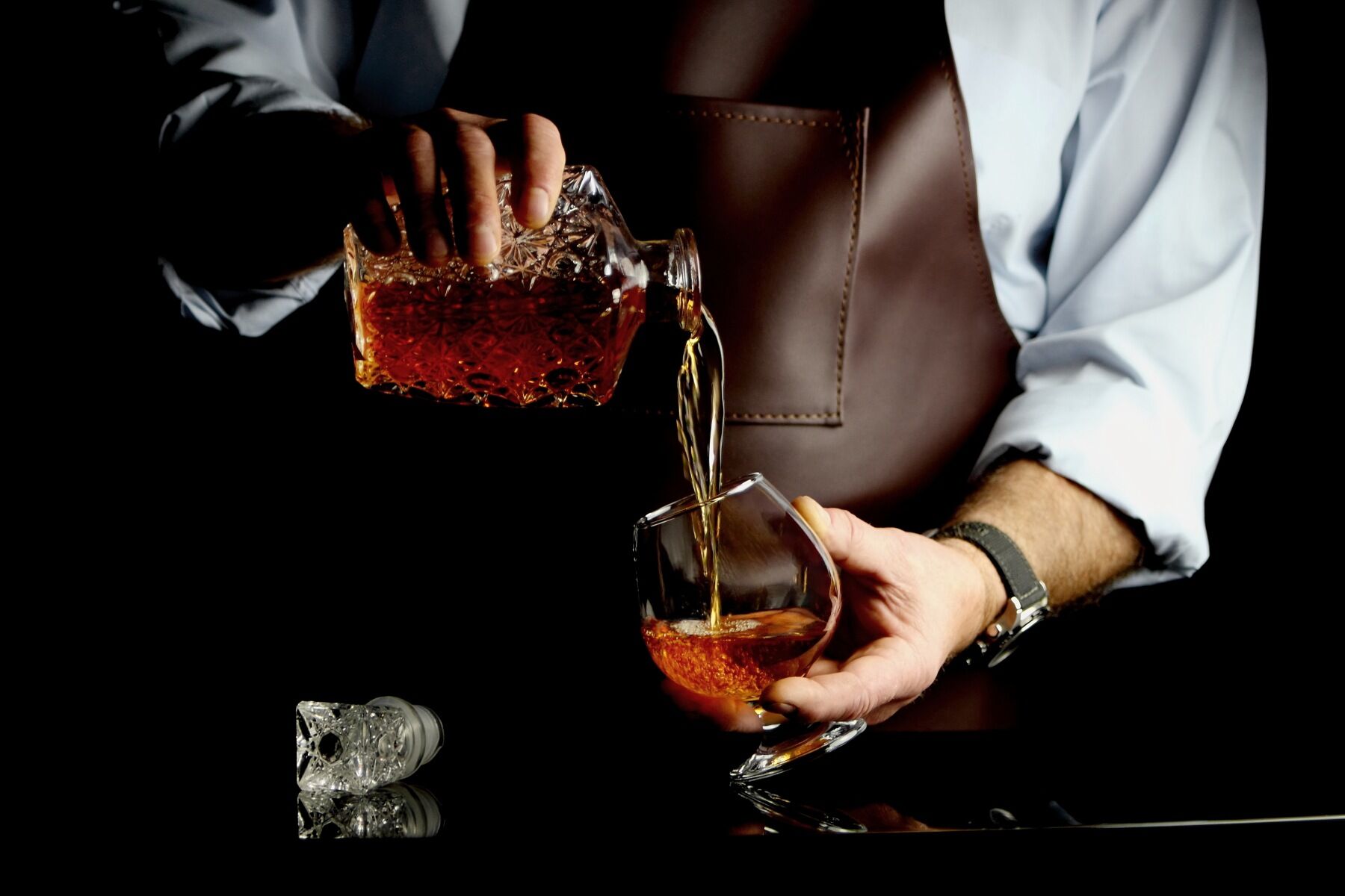 A bartender in a leather apron pouring whisky from a decanter into a glass