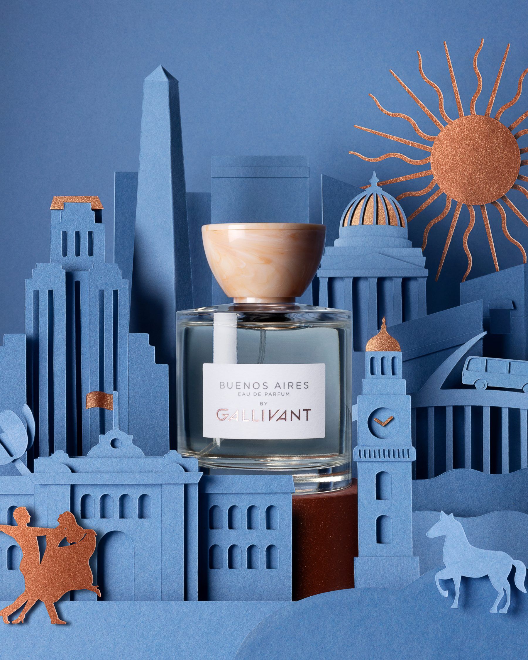 A bottle of Buenos Aires perfume set in front of a diorama depicting Buenos Aires which appears to be made out of blue paper with gold accents